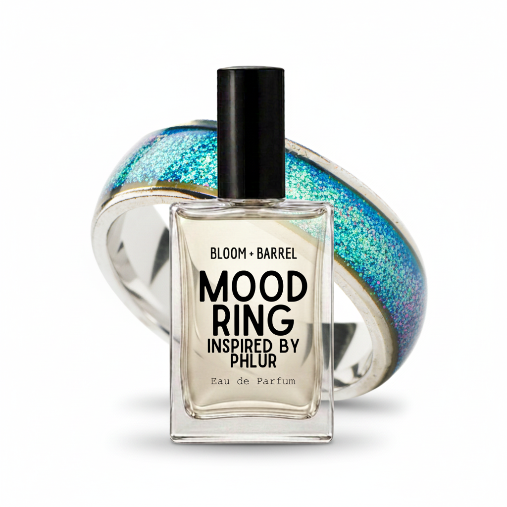 A clear glass perfume bottle labeled 'BLOOM • BARREL MOOD RING INSPIRED BY PHLR Eau de Parfum' with a black cap, set in front of a translucent ring featuring blue, green, and gold glittery segments on a white background.