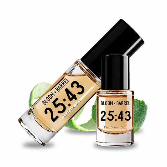 Two clear glass bottles of perfume oil with black caps and black labels reading BLOOM + BARREL 25:43 Perfume Oil, shown on a white background with green lime slices behind them.
