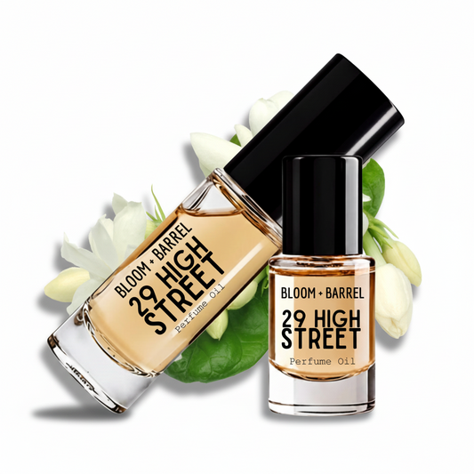 Two glass bottles of Bloom + Barrel 29 High Street perfume oil with black caps, arranged with white flowers and green leaves on a white background.