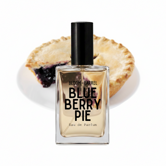A rectangular glass perfume bottle labeled 'BLOOM + BARREL BLUE BERRY PIE Eau de Parfum' with a black cap, shown in front of a sliced blueberry pie on a white background.