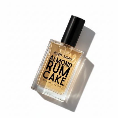 A rectangular glass perfume bottle with almond-shaped edges, filled with amber-colored fragrance liquid and topped with a matte black cap. The front label reads 'BLOOM • BARREL ALMOND RUM CAKE Eau de Parfum' in black text.