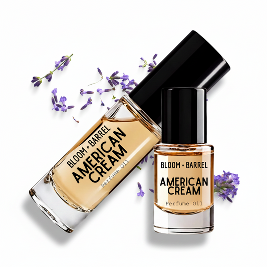 Two clear glass bottles of BLOOM + BARREL American Cream perfume oil with black caps, arranged on a white background with scattered purple lavender sprigs. One bottle is upright and the other is tilted diagonally.