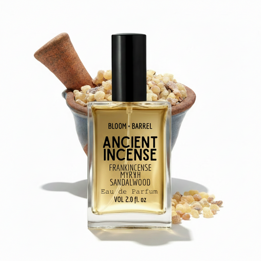 A bottle of Bloom + Barrel Ancient Incense Eau de Parfum, featuring a clear glass bottle with a black cap and golden-amber liquid, set against a white background with a wooden mortar and pestle filled with dried spices.