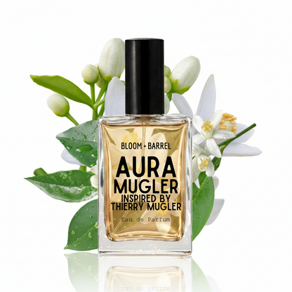 A bottle of Aura Mugler Eau de Parfum with a black cap and amber liquid, styled with white flowers and green leaves on a white background.