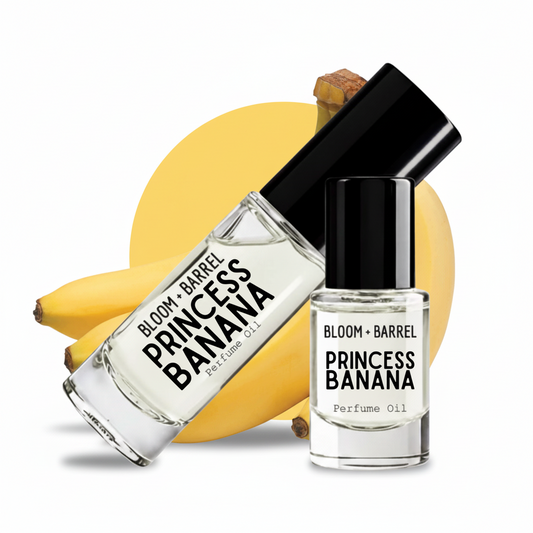 Two bottles of Bloom + Barrel Princess Banana Perfume Oil arranged in front of a bunch of yellow bananas on a white background. One bottle is larger and tilted, and the other is a smaller cylindrical bottle, both with black caps and clear glass showing the pale yellow oil inside.