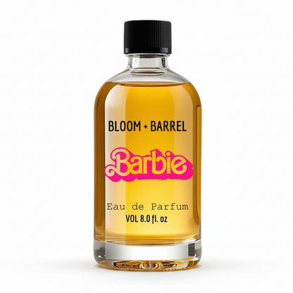 A clear glass bottle of Bloom + Barrel x Barbie Eau de Parfum with golden-yellow liquid, a black cap, and a pink Barbie logo on a white background.