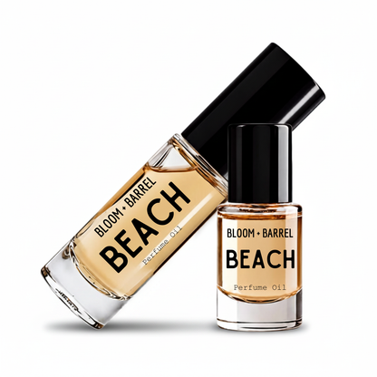 Two clear glass bottles of perfume oil with black caps on a white background. One bottle is tilted diagonally and reads 'BLOOM • BARREL BEACH Perfume Oil', while the other stands upright and reads 'BLOOM • BARREL BEACH' with 'Perfume Oil' below.