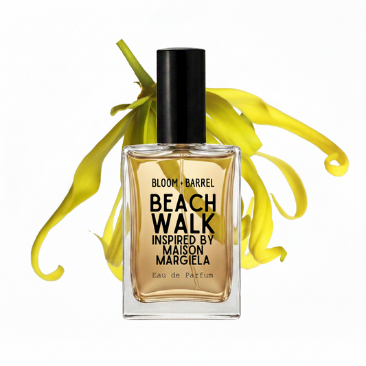 A bottle of Bloom + Barrel Beach Walk Eau de Parfum, inspired by Maison Margiela, featuring amber-colored liquid in a clear glass bottle with a black cap, set against a white background with yellow flower petals.