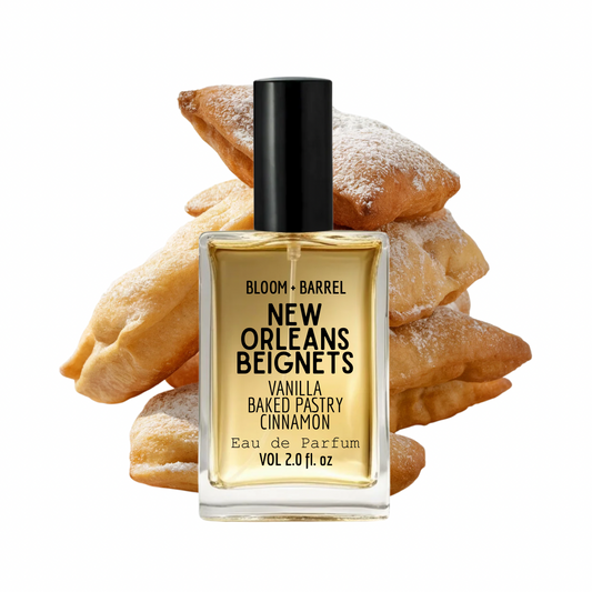A bottle of Bloom + Barrel New Orleans Beignets Eau de Parfum centered in front of a stack of baked beignets on a plain white background.