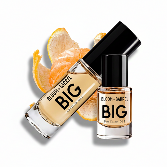 Two glass bottles of BLOOM + BARREL BIG Perfume Oil with black caps, one upright and one tilted, shown with orange peel slices on a white background.