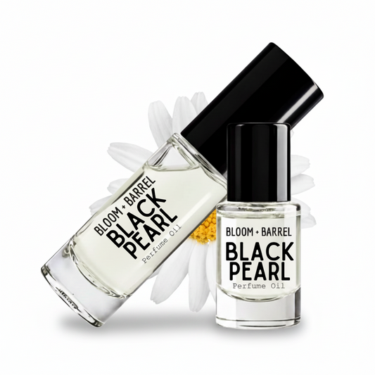 Two glass bottles of BLOOM + BARREL Black Pearl Perfume Oil with black caps, one larger diffuser bottle and one smaller spray bottle, displayed with white daisy petals on a white background.
