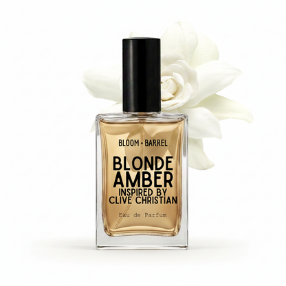 A rectangular glass perfume bottle with a black cap, filled with amber-toned fragrance, labeled 'BLOOM • BARREL BLONDE AMBER INSPIRED BY CLIVE CHRISTIAN Eau de Parfum.' A large white flower is placed behind the bottle on a clean white background.