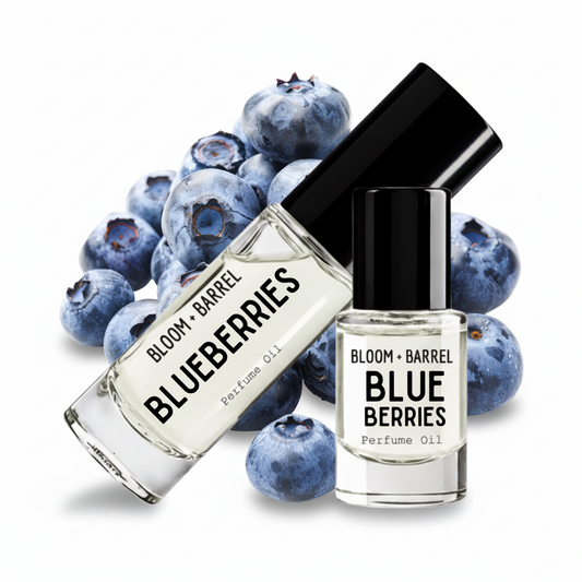 Two glass bottles of Bloom + Barrel Blueberries Perfume Oil with black caps, arranged in front of a pile of fresh blueberries on a white background.