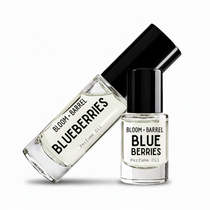 Two clear glass perfume oil bottles with black caps on a white background. One bottle is tilted diagonally and reads 'BLOOM + BARREL BLUEBERRIES Perfume Oil', while the other stands upright with the same label.