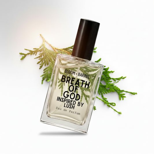 A clear glass perfume bottle with a black cap and black text reading 'BLOOM • BARREL BREATH OF GOD INSPIRED BY LUSH' and 'Eau de Parfum', centered on a white background with green evergreen branches behind it.