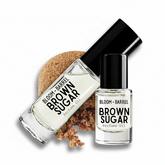 Two bottles of Bloom + Barrel Brown Sugar Perfume Oil on a white background, one larger bottle lying diagonally with brown sugar granules behind it and one smaller upright bottle in front.