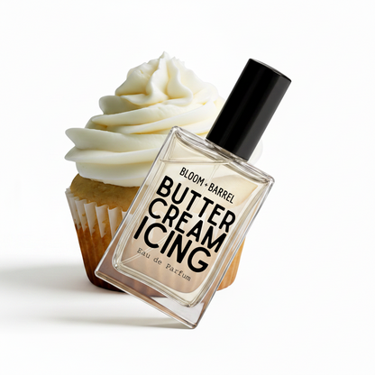 A perfume bottle labeled 'BLOOM • BARREL BUTTER CREAM ICING' with a black cap, shown in front of a cupcake with white frosting on a plain white background.