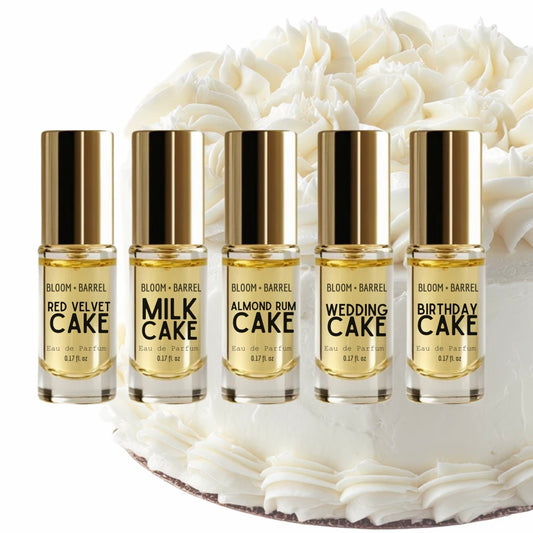 Five small Bloom + Barrel perfume oil bottles with gold caps are lined up in front of a white frosted cake. The bottles are labeled Red Velvet Cake, Milk Cake, Almond Cake, Wedding Cake, and Birthday Cake.