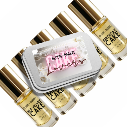 A set of Bloom Barrel perfume bottles with gold caps arranged around a silver rectangular tin labeled 'Cake Lovers' on a white background.