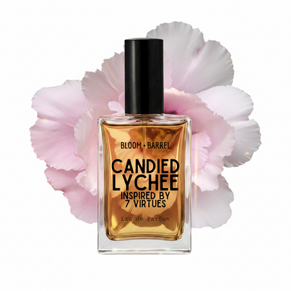A rectangular glass perfume bottle with amber liquid and a black cap, centered in front of large pale pink flower petals on a white background. The bottle label reads 'BLOOM • BARREL', 'CANDIED LYCHEE', and 'INSPIRED BY 7 VIRTUES'.