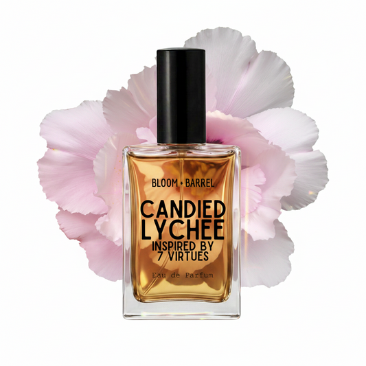A rectangular glass perfume bottle with amber liquid and a black cap, centered in front of large pale pink flower petals on a white background. The bottle label reads 'BLOOM • BARREL', 'CANDIED LYCHEE', and 'INSPIRED BY 7 VIRTUES'.