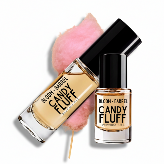 Two bottles of Bloom + Barrel Candy Fluff perfume oil on a white background, one upright and one tilted, with a pink cotton candy graphic behind them and a wooden stick holding the tilted bottle.