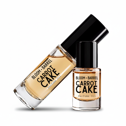 Two glass bottles of BLOOM + BARREL carrot cake Perfume Oil with black caps, one larger bottle lying diagonally and one smaller bottle standing upright, on a white background.