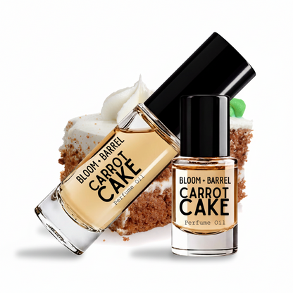 Two bottles of Bloom + Barrel Carrot Cake Perfume Oil arranged on a white background with a carrot cake slice behind them. One bottle is tilted diagonally and the other stands upright, both showing the product name and brand on the labels.