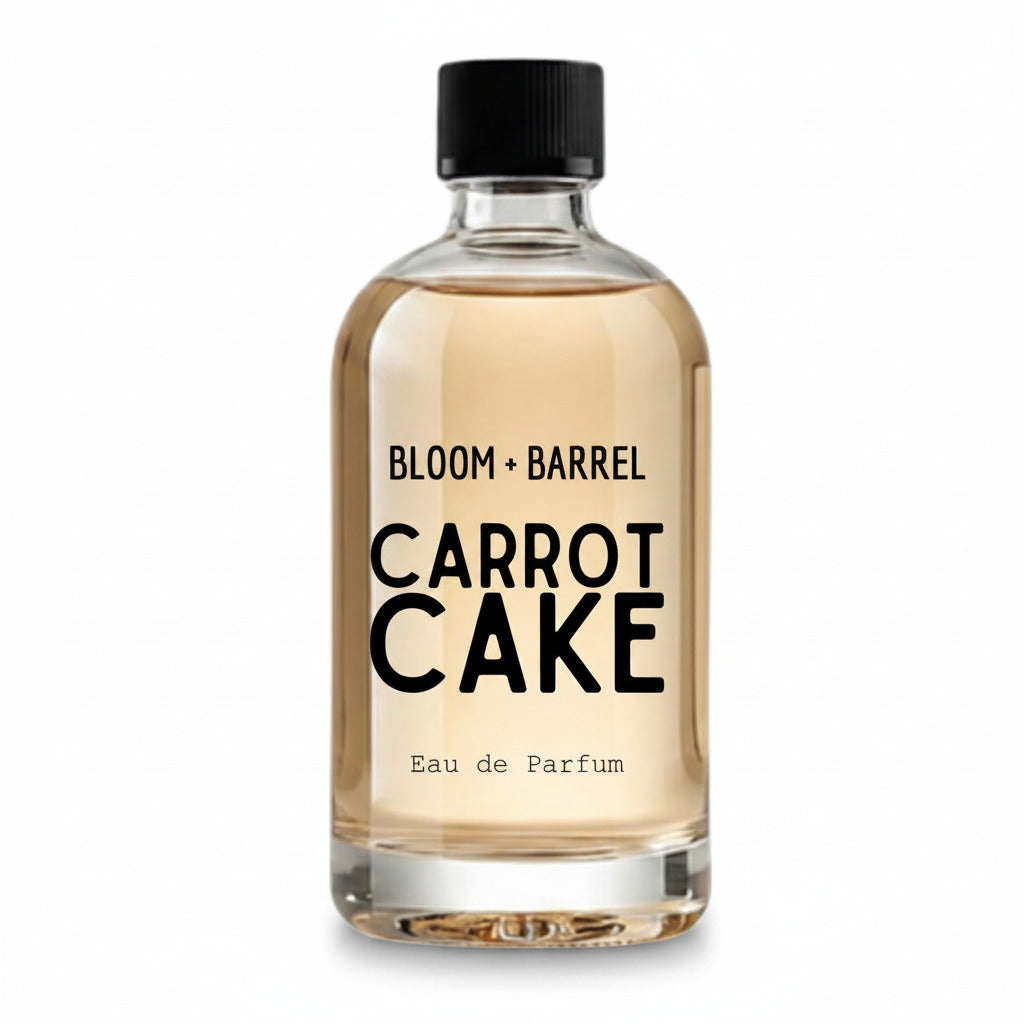A clear glass bottle of Bloom + Barrel Carrot Cake Eau de Parfum with a black cap, filled with a light golden-amber liquid, on a solid white background.