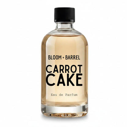 A clear glass bottle of Bloom + Barrel Carrot Cake Eau de Parfum with a black cap, filled with a light golden-amber liquid, on a solid white background.