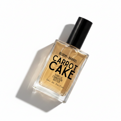 A rectangular glass perfume bottle with a black cap, filled with a warm amber liquid. The front label reads 'BLOOM • BARREL CARROT CAKE VANILLA CINNAMON BUTTER Eau de Parfum' in black text, set against a plain white background.