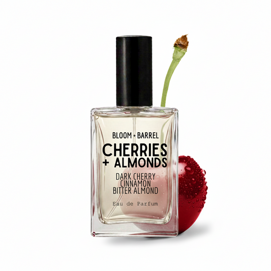 A rectangular glass perfume bottle labeled 'BLOOM + BARREL CHERRIES + ALMONDS' with a black cap, shown on a white background with a cherry stem and a red cherry beside it.