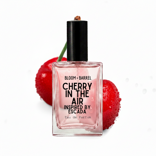 A rectangular glass perfume bottle labeled 'BLOOM • BARREL CHERRY IN THE AIR INSPIRED BY ESCADA Eau de Parfum' with a black cap, centered in front of two glossy red cherries on a white background.