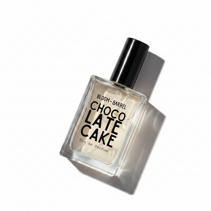 A rectangular glass perfume bottle with a black cap, containing a pale golden liquid. The front label reads 'BLOOM • BARREL CHOCOLATE CAKE Eau de Parfum' in black text, set against a plain white background.