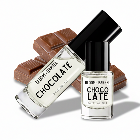 Two clear perfume oil bottles labeled BLOOM + BARREL CHOCOLATE, one with a black cap standing upright and one tilted diagonally, set against brown chocolate pieces on a white background.