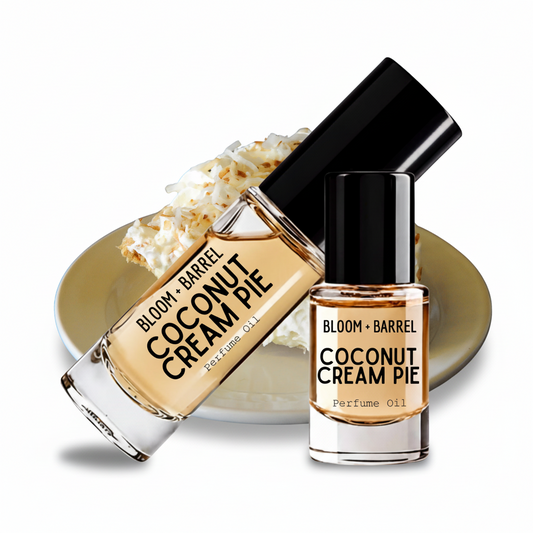 Two glass bottles of BLOOM + BARREL Coconut Cream Pie Perfume Oil with black caps, one upright and one tilted, displayed on a gold plate with whipped cream against a white background.