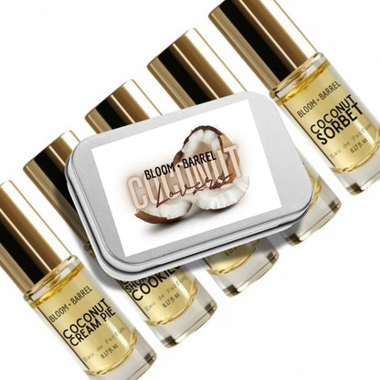 A set of Bloom+ Barrel coconut-scented perfume or fragrance bottles with gold caps arranged around a small rectangular tin labeled 'Coconut' and showing coconut halves. The bottles contain a pale golden liquid and are photographed on a clean white background.