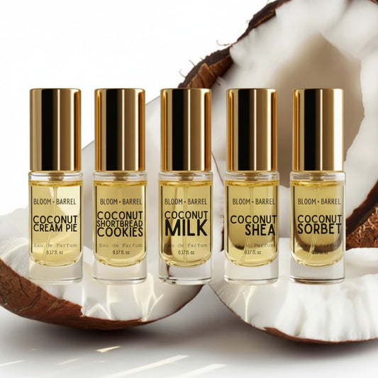 Five Bloom + Barrel coconut-themed perfume rollerball bottles arranged in a row on a white surface with halved coconuts behind them. The bottles contain pale yellow liquid and have shiny gold caps.