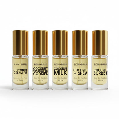 Five small clear glass bottles with metallic gold caps and gold-toned liquid, arranged in a row on a white background. The labels read Bloom + Barrel and various coconut-flavored variants including Cream Pie, Shortbread Cookies, Milk, + Shea, and Sorbet.