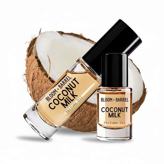 Two glass bottles of Bloom + Barrel Coconut Milk Perfume Oil with black caps, one larger bottle tilted over a sliced coconut and one smaller bottle, on a white background.