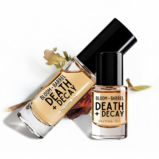 Two glass perfume oil bottles with black caps on a white background, one upright and one tilted, with dried leaves and a cinnamon stick as styling props. The bottles are labeled BLOOM + BARREL DEATH + DECAY Perfume Oil.