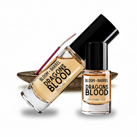 Two glass bottles of Bloom + Barrel Dragons Blood Perfume Oil with black caps, one larger diffuser-style bottle and one smaller perfume oil bottle, shown on a white background with a small dish behind them.