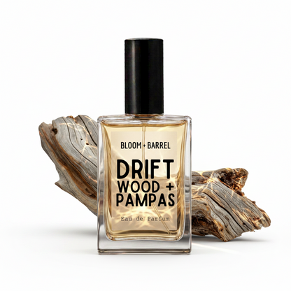 A rectangular glass perfume bottle with a black cap, labeled 'BLOOM • BARREL DRIFT WOOD + PAMPAS Eau de Parfum', centered against a white background with pieces of weathered driftwood behind it.