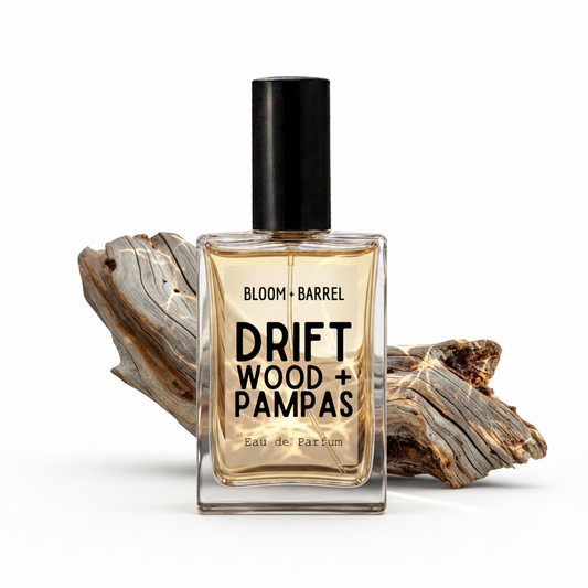 A rectangular glass perfume bottle with a black cap, labeled 'BLOOM • BARREL DRIFT WOOD + PAMPAS Eau de Parfum', centered against a white background with pieces of weathered driftwood behind it.
