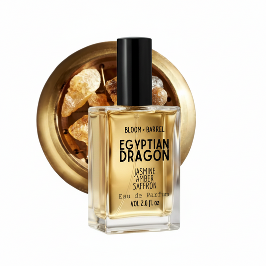 A bottle of Bloom + Barrel Egyptian Dragon Jasmine Amber Saffron Eau de Parfum, featuring a clear glass bottle with golden-yellow liquid, a black cap, and a gold-toned circular background graphic containing amber crystals.