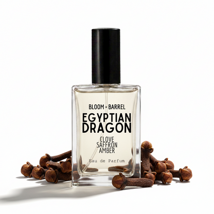 A rectangular glass perfume bottle labeled 'BLOOM + BARREL EGYPTIAN DRAGON CLOVE SAFFRON AMBER' with a black cap, centered on a white background and framed by cloves on both sides.