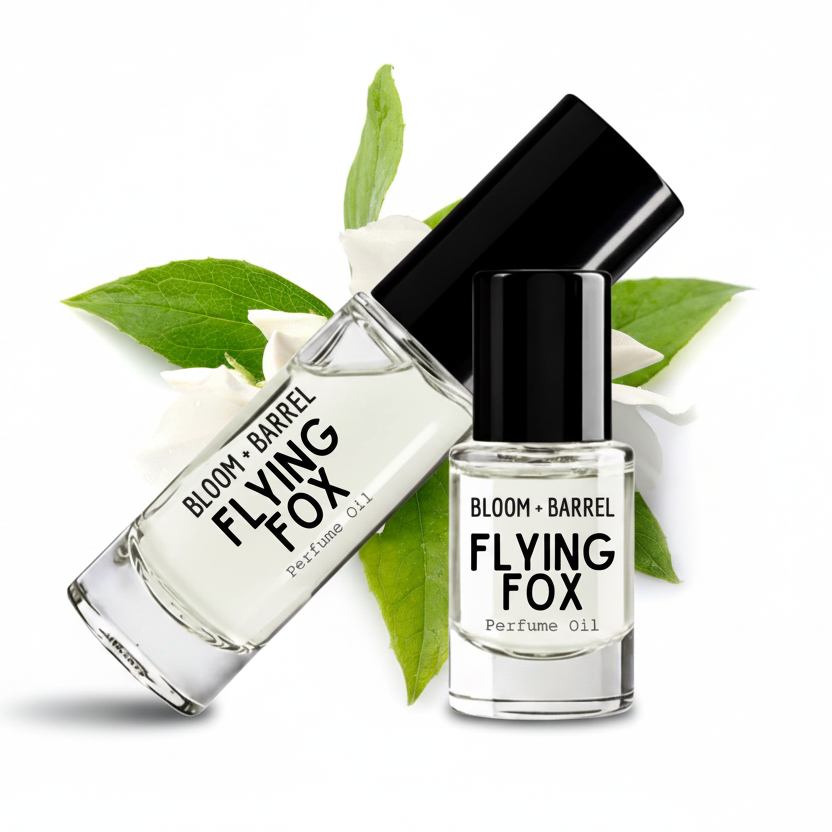 Two clear glass bottles of Bloom + Barrel Flying Fox perfume oil with black caps, arranged on a white background with green leaves and white blossoms.