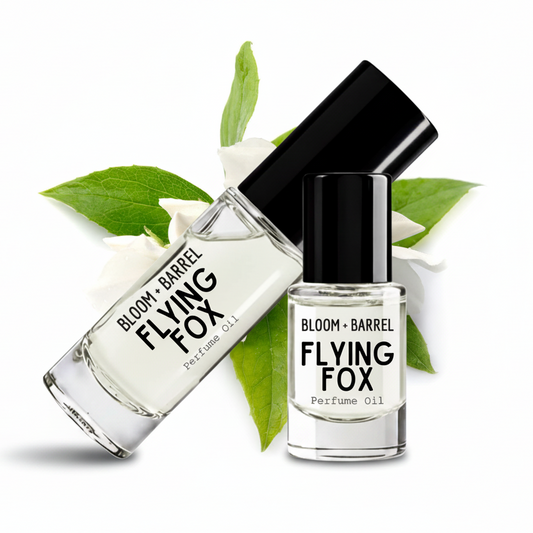 Two clear glass bottles of Bloom + Barrel Flying Fox perfume oil with black caps, arranged on a white background with green leaves and white blossoms.