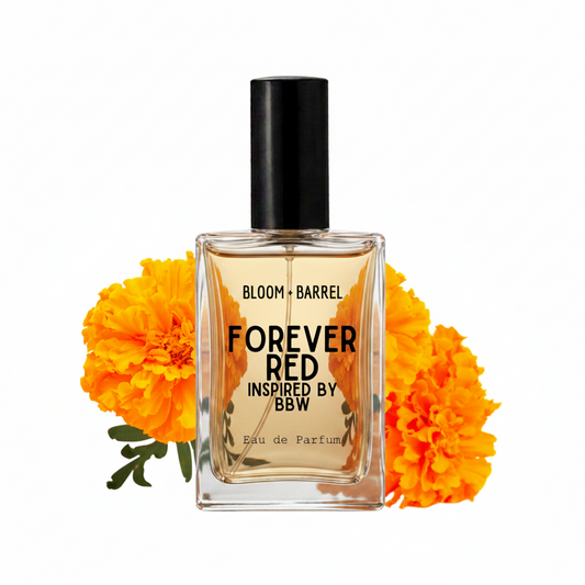 A rectangular glass perfume bottle with a black cap, labeled 'FOREVER RED INSPIRED BY BBW' and 'BLOOM • BARREL', centered on a white background with bright orange marigold flowers behind it.