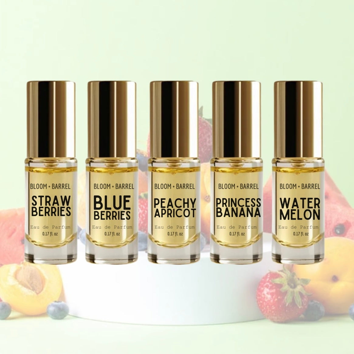 Five small Bloom+Barrel perfume bottles with metallic gold caps are lined up on a white pedestal. The bottles contain pale yellow liquid and are labeled with different scent names: Strawberry, Blue Berries, Peachy Apricot, Princess Banana, and Watermelon.